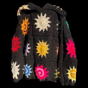 May include: A black crochet hoodie with vibrant sunburst motifs in yellow, orange, pink, red, and white. The hoodie includes a hood and is crafted from a textured, handmade material. The sunbursts are square and regularly spaced.