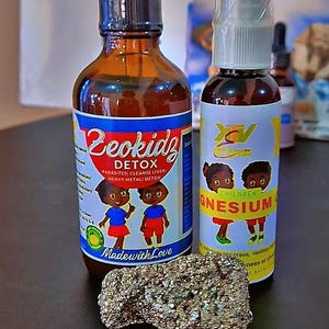 May include: Two brown glass bottles with labels, one labeled "Zeokidz Detox" and the other "Children Magnesium", are displayed. A rough, metallic-looking rock sits in front of the bottles. The bottles have cartoon illustrations.