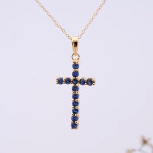 Puede incluir: Un collar con colgante dorado con un diseño de cruz. La cruz está adornada con numerosas piedras preciosas pequeñas, redondas y de color azul oscuro. La cadena del collar es delicada y también dorada, creando un aspecto elegante y cohesivo.