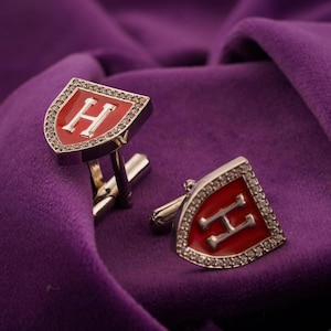 May include: A pair of silver cufflinks with a red enamel shield design. Each cufflink features a white letter "H" and is bordered with small, clear crystals. The cufflinks are displayed on a purple velvet surface.