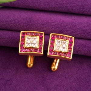 Peut inclure: Une paire de boutons de manchette dorés de forme carrée. Chaque bouton de manchette présente un carré central de pierres blanches, entouré d'une bordure de pierres roses. Les boutons de manchette sont posés sur un fond violet.