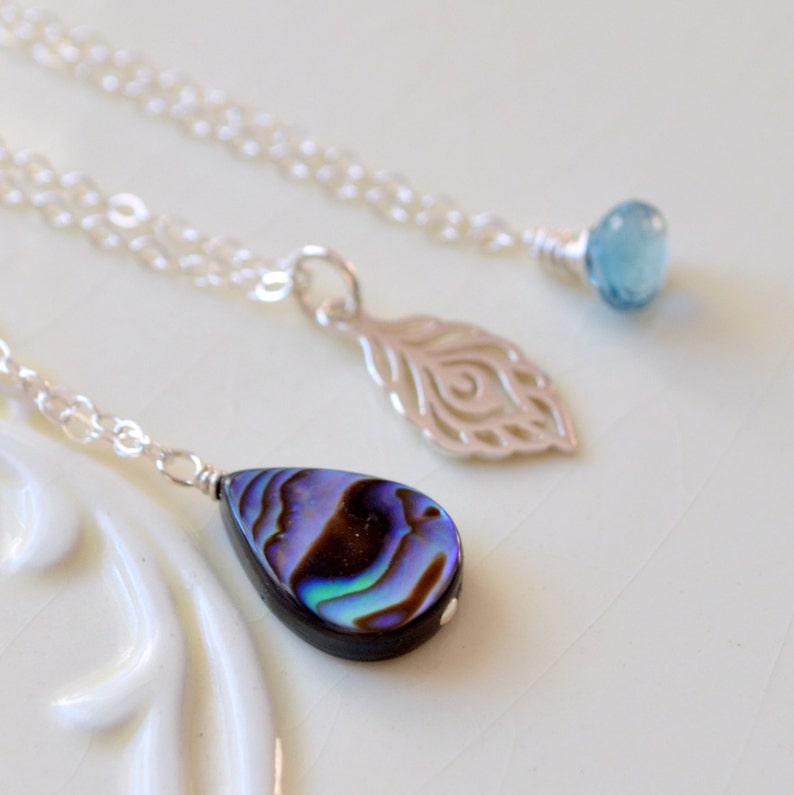 May include: Three silver necklaces with different pendants. The first necklace has a light blue gemstone pendant. The second necklace has a silver peacock feather pendant. The third necklace has a teardrop-shaped iridescent abalone shell pendant.