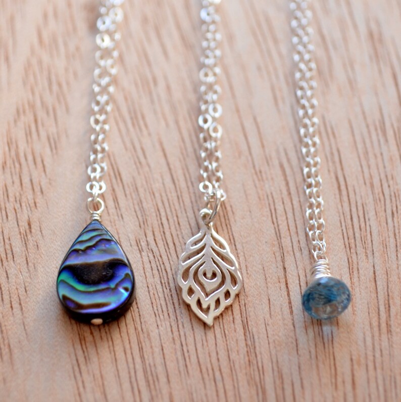 May include: Three silver necklaces with different pendants. The first necklace has a teardrop-shaped pendant made of iridescent abalone shell. The second necklace has a delicate silver pendant shaped like a feather. The third necklace has a small, round, light blue gemstone pendant.