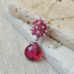May include: A silver necklace featuring a pendant with a cluster of small, pink gemstones and a large, faceted, teardrop-shaped ruby. The pendant is suspended from a delicate silver chain, showcasing the vibrant red and pink hues of the gemstones.