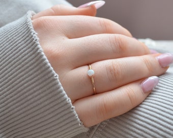 May include: A delicate gold ring with a small white opal gemstone. The ring is worn on a finger with pink nail polish.