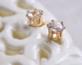 May include: A pair of gold stud earrings with clear round gemstones set in a crown-like design.