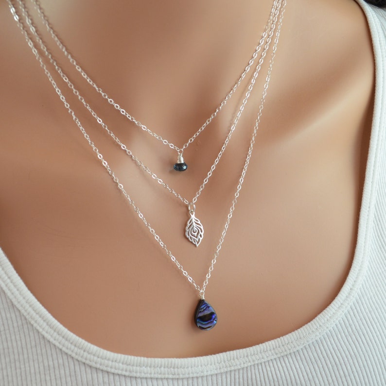 May include: A layered silver necklace with three pendants. The top pendant is a small, dark blue gemstone. The middle pendant is a silver leaf design. The bottom pendant is a teardrop-shaped, iridescent blue gemstone.