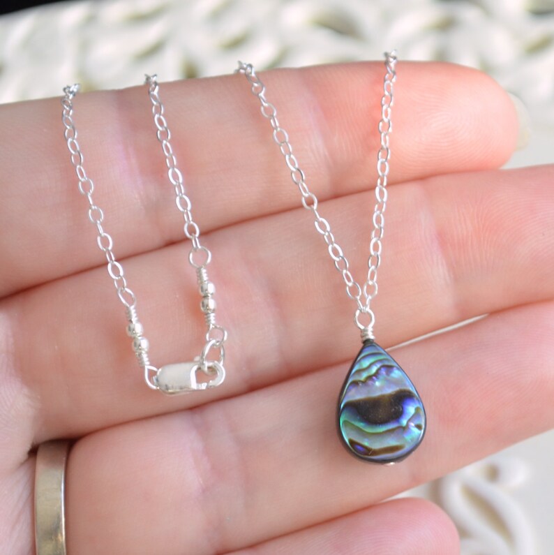 May include: A silver chain necklace with a teardrop-shaped abalone shell pendant. The shell has iridescent blue, green, and black colors.
