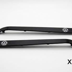 Puede incluir: Dos piezas de moldura automotriz negras, alargadas y estrechas, con una ligera curva en un extremo. Cada pieza presenta un emblema circular blanco y negro con el logotipo de Volkswagen cerca del extremo. El texto x2 es visible abajo a la derecha.
