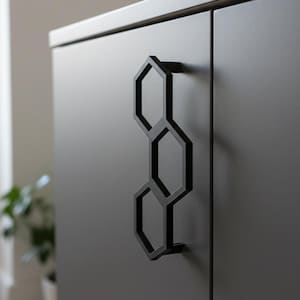 Peut inclure: Un meuble gris foncé avec une poignée géométrique noire. La poignée est composée de trois formes hexagonales reliées. Le meuble a une finition lisse. La poignée est montée verticalement sur la porte.