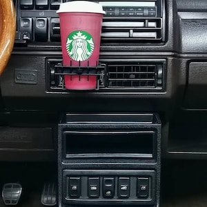 May include: A red Starbucks coffee cup with a white lid sits in a black car vent cup holder. The Starbucks logo is displayed on the cup. The car's dashboard is dark grey with various buttons and vents.