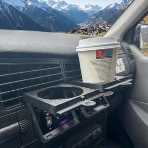 Op de afbeelding: Zwarte 3D-geprinte bekerhouder en dienblad voor in de auto, gemonteerd op het dashboard. Een witte papieren koffiebeker staat in het dienblad. Het interieur van de auto is zichtbaar, met een berglandschap op de achtergrond.