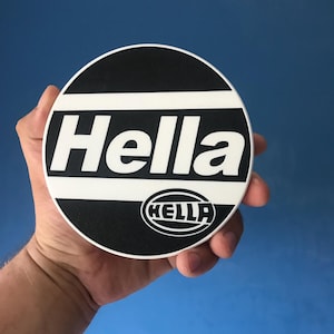 Pode incluir: Um porta-copos circular preto e branco com a palavra Hella em letras brancas grandes no centro, com um logótipo Hella mais pequeno numa forma oval posicionado na parte inferior direita do design sobre um fundo preto.