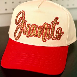 May include: A cream-colored baseball cap with a bright red brim and a small red button on top. The front of the cap features the name Juanito written in a cursive script, filled with gold-toned rhinestones and outlined in red glitter.