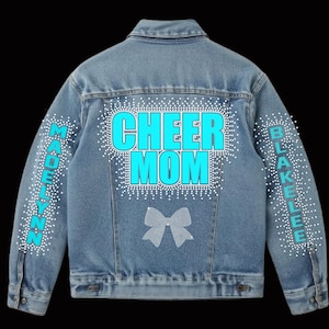 May include: Light blue denim jacket with the words "CHEER MOM" in large, turquoise letters on the back, surrounded by white rhinestones. The sleeves have the names "MADELYNN" and "BLAKELEE" in turquoise. A rhinestone bow is below the text.