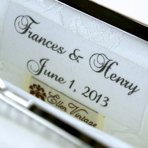 Può includere: Primo piano dell'interno di una pochette in raso bianco con i nomi "Frances & Henry" e la data "1 giugno 2013" stampati in corsivo nero. Una piccola etichetta recita "Ellen Vintage". È visibile una chiusura argentata.