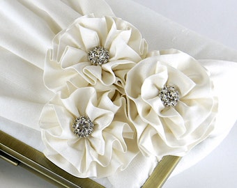May include: A white satin clutch bag with three large white fabric flowers adorned with sparkling rhinestones.