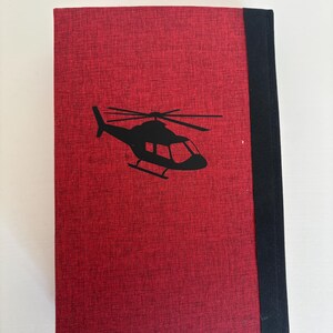 May include: A red book with a black helicopter silhouette on the cover. The book has a textured surface and a black spine. The helicopter design is simple and modern.