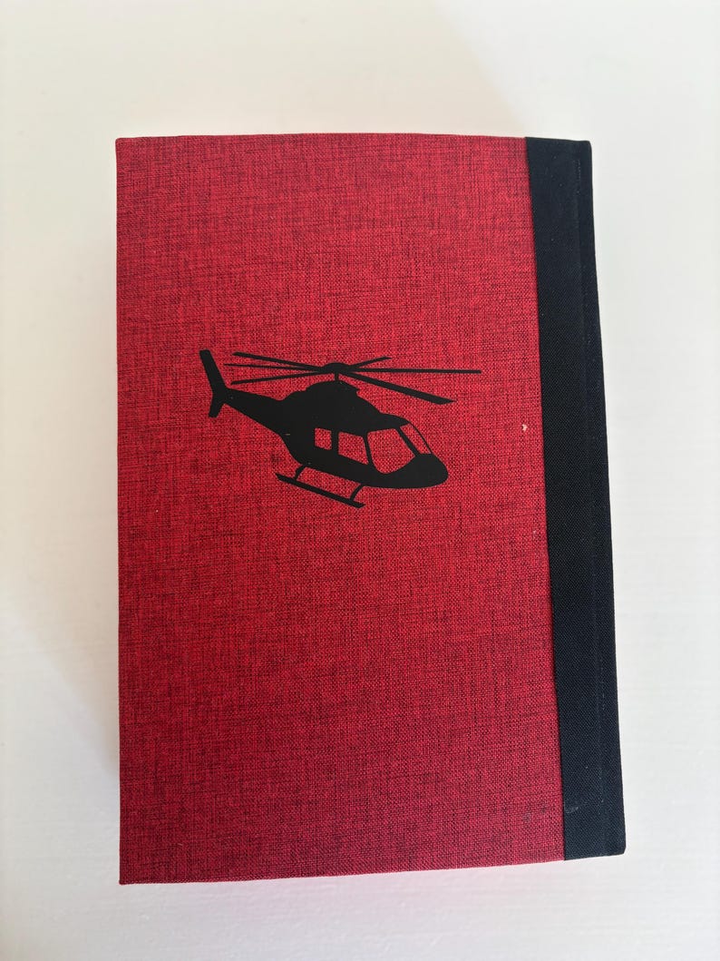 May include: A red book with a black helicopter silhouette on the cover. The book has a textured surface and a black spine. The helicopter design is simple and modern.