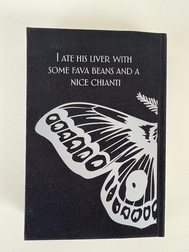 May include: A black book cover with the text "I ATE HIS LIVER WITH SOME FAVA BEANS AND A NICE CHIANTI" in white. A large, stylized butterfly wing design in white is featured on the lower portion of the cover.