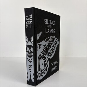 May include: A black book titled "Silence of the Lambs" by Thomas Harris. The cover features a large silver moth and the title in silver lettering. The spine displays the title and a skull design.