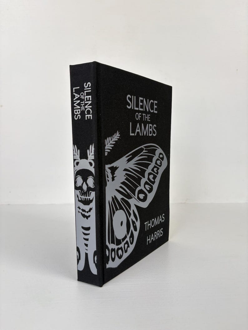 May include: A black book titled "Silence of the Lambs" by Thomas Harris. The cover features a large silver moth and the title in silver lettering. The spine displays the title and a skull design.