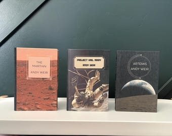 Puede incluir: Tres libros de tapa dura de Andy Weir dispuestos uno al lado del otro. El primer libro se titula The Martian con una portada de paisaje rojo. El segundo libro es Project Hail Mary con un astronauta. El tercer libro es Artemis con una vista lunar.