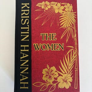 May include: A hardcover book titled "THE WOMEN" by Kristin Hannah. The book has a red cover with gold floral illustrations and text. The spine is black with gold lettering.