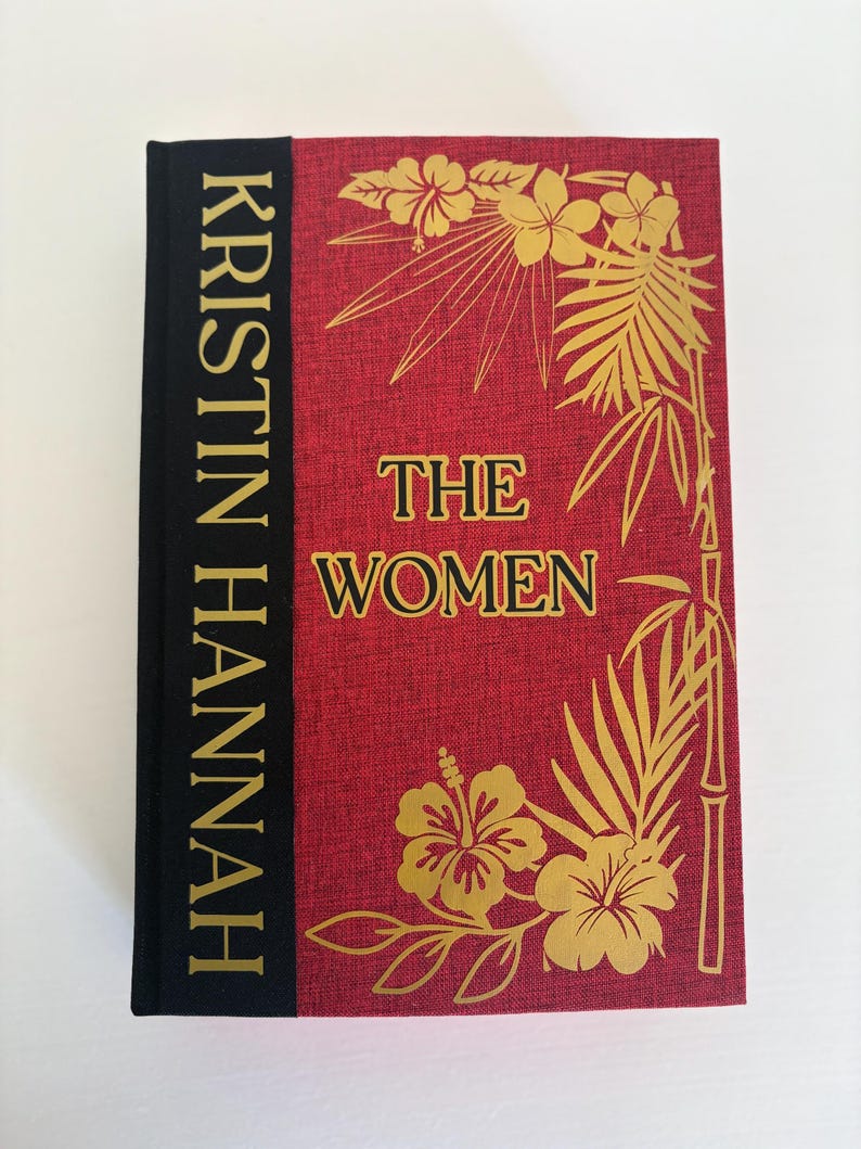 May include: A hardcover book titled "THE WOMEN" by Kristin Hannah. The book has a red cover with gold floral illustrations and text. The spine is black with gold lettering.