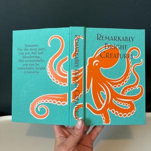 May include: An open book with a teal cover featuring an orange octopus illustration and the title "Remarkably Bright Creatures." The book's spine also displays the title. Text on the left page reads, "Humans. For the most part, you are dull and blundering. But occasionally, you can be remarkably bright creatures."