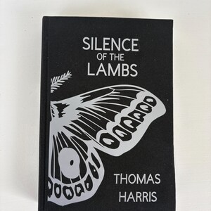 May include: A hardcover book titled "SILENCE OF THE LAMBS" by Thomas Harris. The book has a black cover with a large, stylized moth graphic in white. The title and author's name are also in white.