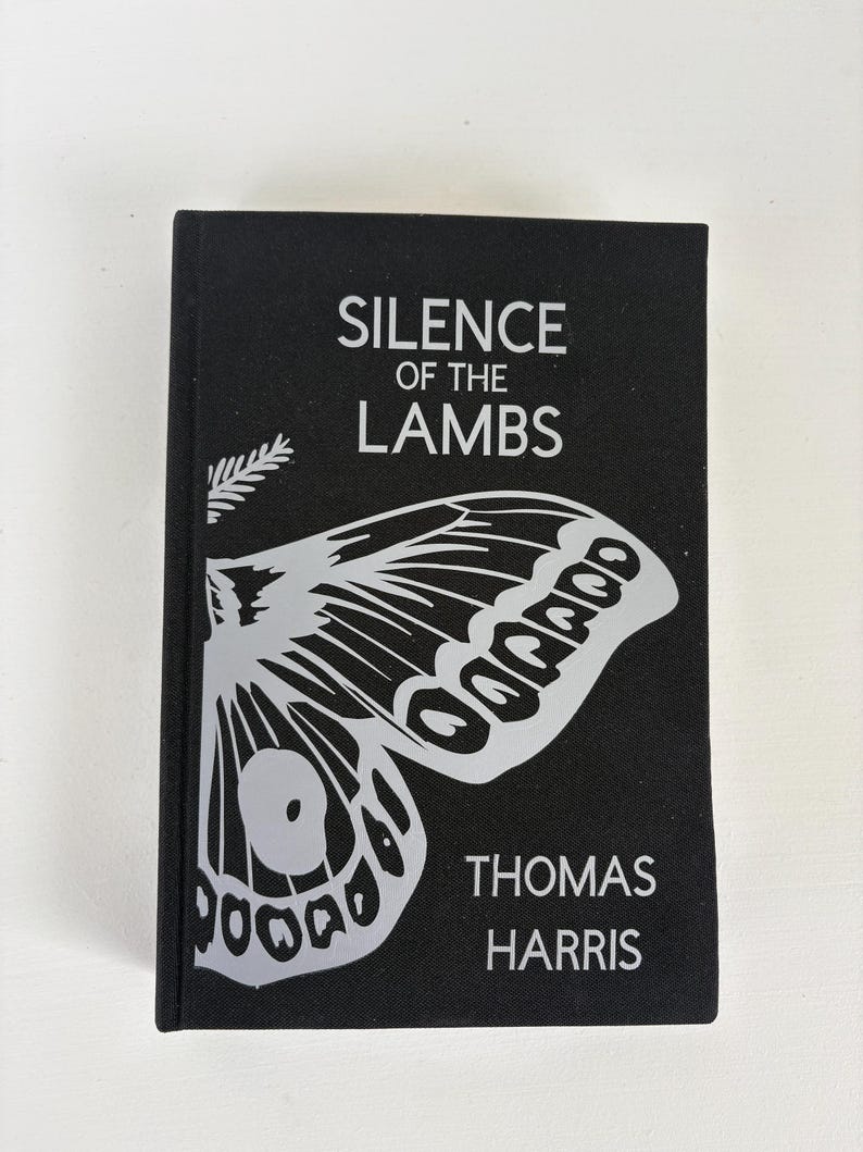 May include: A hardcover book titled "SILENCE OF THE LAMBS" by Thomas Harris. The book has a black cover with a large, stylized moth graphic in white. The title and author's name are also in white.