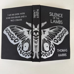 May include: A hardcover book titled "Silence of the Lambs" by Thomas Harris. The black cover features a silver moth design with a skull and the text "I ate his liver with some fava beans and a nice chianti."