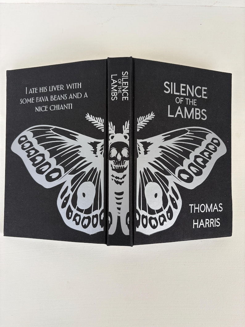 May include: A hardcover book titled "Silence of the Lambs" by Thomas Harris. The black cover features a silver moth design with a skull and the text "I ate his liver with some fava beans and a nice chianti."