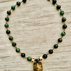 May include: A beaded necklace featuring dark green and black beads with a gold-colored turtle pendant. The necklace has a toggle clasp and includes small white beads with star designs.