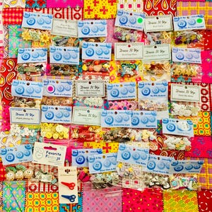 May include: A colorful patchwork background displays various craft supplies. Packages of buttons, embellishments, and small scissors are visible. The buttons come in various colors and designs, perfect for sewing and crafting projects.