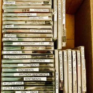 May include: A cardboard box filled with clear plastic cases, each labeled with various words like "Christmas," "Sentiments," and "Friends." The cases appear to contain craft supplies or embellishments, with a variety of fonts and styles.