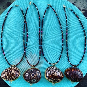 May include: Four necklaces with shell pendants. The pendants have a brown and cream spotted pattern, with gold-colored metal trim. The necklaces have black beaded strands with colorful accents. The necklaces are displayed on a turquoise blue textured surface.