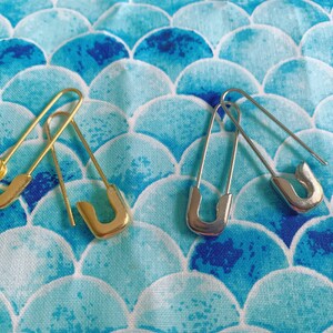 Puede incluir: Dos imperdibles, uno dorado y otro plateado, se muestran sobre una tela estampada en azul y blanco. Los imperdibles est&aacute;n abiertos, mostrando su dise&ntilde;o cl&aacute;sico. La tela tiene un patr&oacute;n abstracto repetitivo.