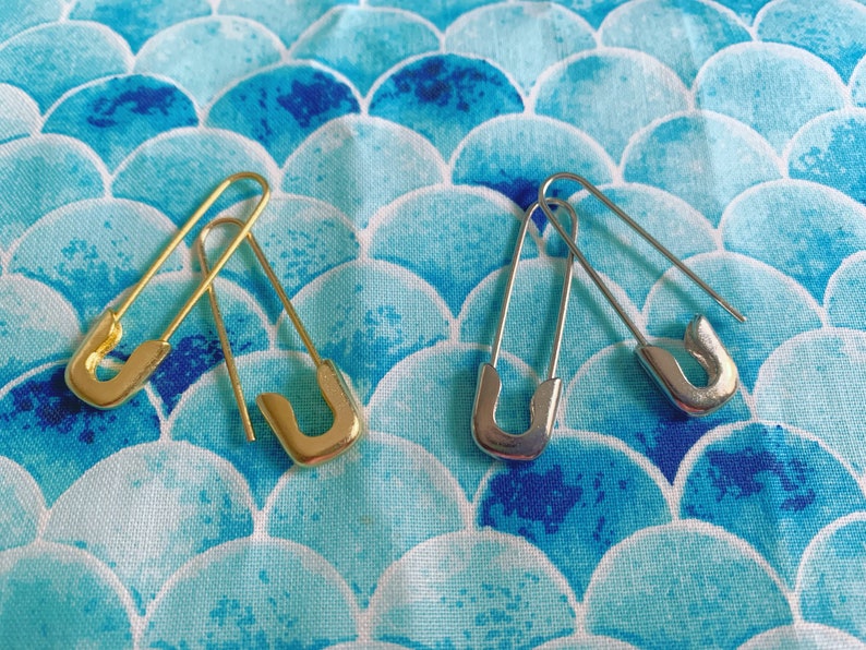 Puede incluir: Dos imperdibles, uno dorado y otro plateado, se muestran sobre una tela estampada en azul y blanco. Los imperdibles est&aacute;n abiertos, mostrando su dise&ntilde;o cl&aacute;sico. La tela tiene un patr&oacute;n abstracto repetitivo.
