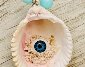 May include: A unique necklace featuring a seashell pendant with a blue eye and sand accents. Two light blue beads are attached to the top of the shell. The shell is white and pink, with a clear string.