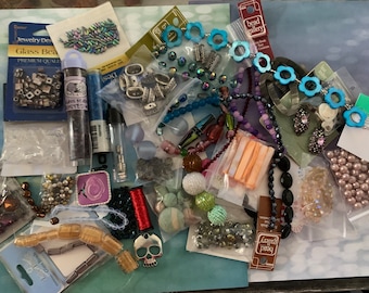 May include: A collection of jewelry-making supplies, including various beads in different shapes, sizes, and colors. There are glass beads, seed beads, and other decorative elements. Some items are packaged, and others are loose. The color palette includes blues, purples, and iridescent tones.