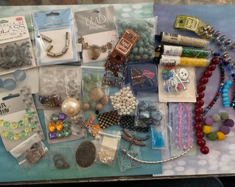 May include: A collection of various beads and jewelry-making supplies. The image includes bags of glass beads, faux pearls, and decorative elements like dragonfly charms and skull-and-crossbones beads. Colors range from clear and iridescent to vibrant hues.