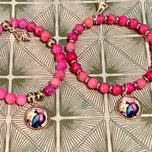 May include: Two pink beaded bracelets with silver accents and round pendants featuring a colorful bird design. The bracelets are made of pink beads and silver-colored metal. The background is a green and white geometric pattern.