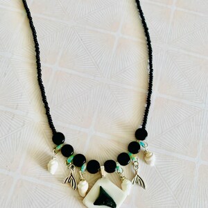 May include: A necklace featuring a black beaded chain with a central white diamond-shaped pendant containing a dark shark tooth. The design includes silver-toned metal mermaid tail charms and small white seashell accents interspersed with black beads.
