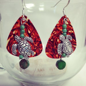 May include: A pair of dangle earrings featuring a red and orange patterned teardrop-shaped pendant. Each earring has a silver-colored turtle charm, green beads, and a green stone at the bottom. The earrings hang from silver-colored hooks.