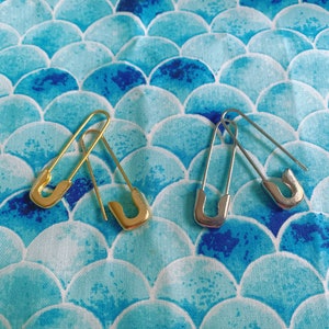 Puede incluir: Dos imperdibles, uno dorado y otro plateado, descansan sobre una tela azul y blanca con un patr&oacute;n de escamas. Los imperdibles est&aacute;n abiertos, mostrando su dise&ntilde;o simple y funcionalidad. El patr&oacute;n de la tela a&ntilde;ade un elemento decorativo.