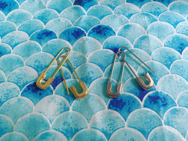 Puede incluir: Dos imperdibles, uno dorado y otro plateado, descansan sobre una tela azul y blanca con un patr&oacute;n de escamas. Los imperdibles est&aacute;n abiertos, mostrando su dise&ntilde;o simple y funcionalidad. El patr&oacute;n de la tela a&ntilde;ade un elemento decorativo.