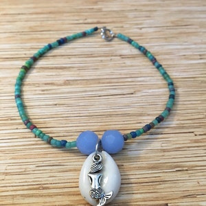 May include: A beaded necklace with a mermaid pendant. The necklace features a strand of small, colorful beads in shades of green, blue, and brown. Two light blue beads flank a white shell pendant with a silver mermaid charm.