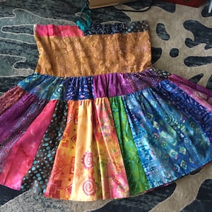 May include: A colorful, handmade skirt with a patchwork design. The skirt features a variety of vibrant fabrics in shades of pink, purple, orange, blue, and green. The skirt has a wide, gathered skirt and a fitted bodice. The skirt is likely made of cotton.